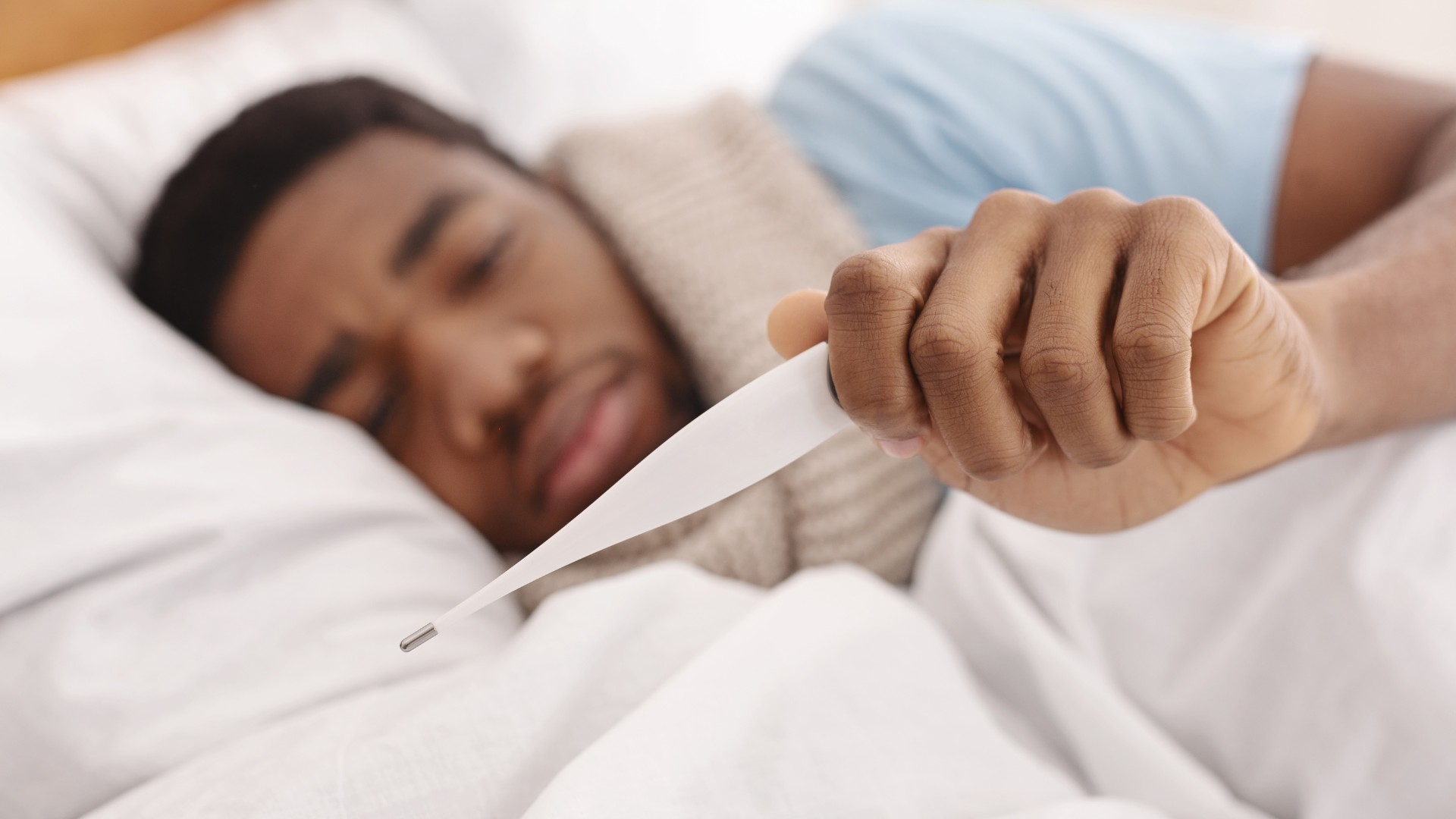 A man lying in bed holding a thermometer, representing fever, an early HIV symptom