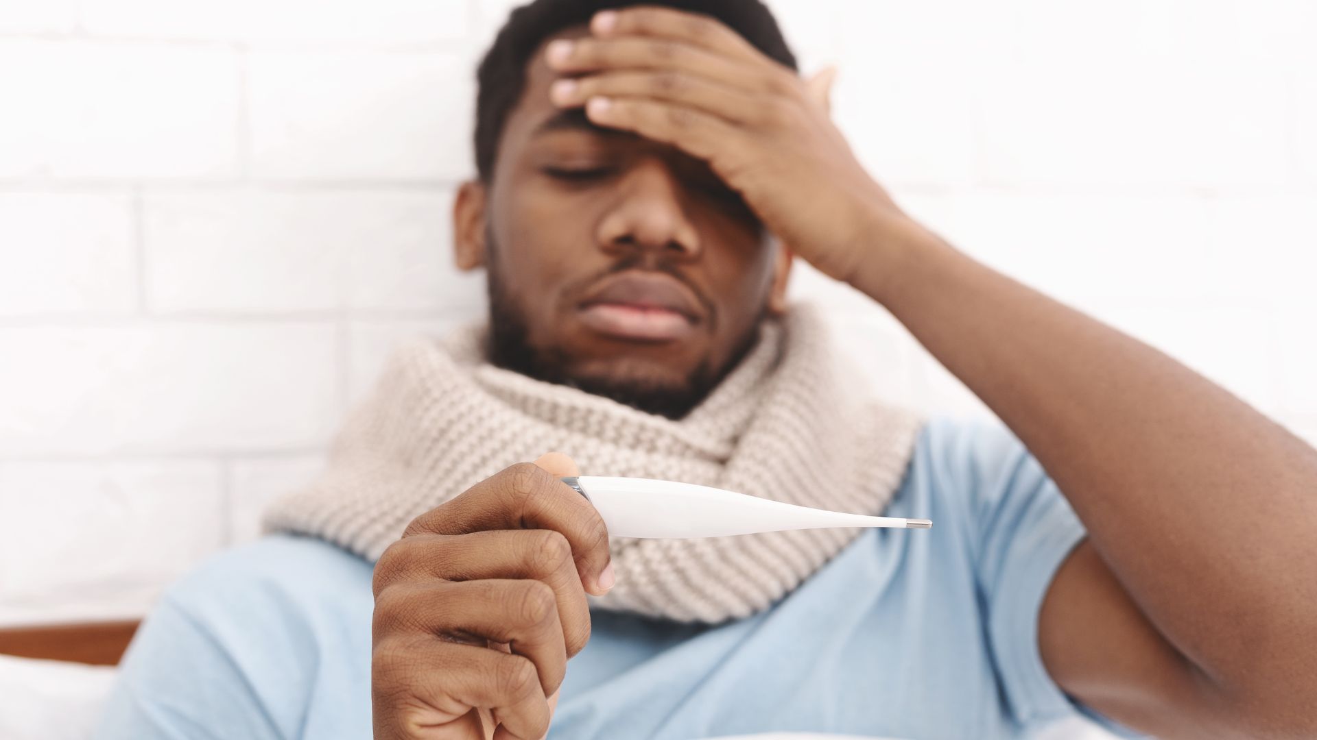 A man sitting up in bed with a hand on his forehead, checking a thermometer for fever
